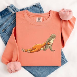 Illustration of a baby macaque named Punch holding a small toy macaque close to his chest, showing an emotional and gentle pose inspired by the viral story of the abandoned monkey, with the larger macaque lying down and the smaller toy being hugged tightly, rendered in a detailed DTF print style with a soft knitted and patch-like stitched effect, featuring clean outlines and layered artwork that gives the design a textured.