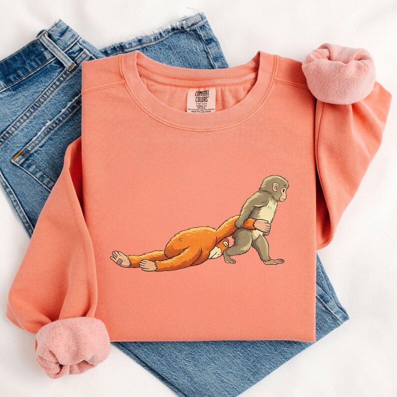 Illustration of a baby macaque named Punch holding a small toy macaque close to his chest, showing an emotional and gentle pose inspired by the viral story of the abandoned monkey, with the larger macaque lying down and the smaller toy being hugged tightly, rendered in a detailed DTF print style with a soft knitted and patch-like stitched effect, featuring clean outlines and layered artwork that gives the design a textured.