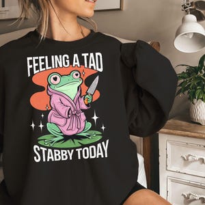 May include: Black sweatshirt featuring a cartoon frog in a pink robe holding a knife. The frog is on a lily pad with the text "FEELING A TAD STABBY TODAY" in white. The design includes orange, green, and white accents.
