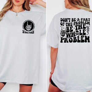 T-shirt featuring a front chest circle design with a skeleton hand and the words “The Whole Problem” surrounding it; the back showcases a large statement reading “Don’t Be a Part of the Problem, Be the Whole Problem” in bold font, with graphic accents like lightning bolts, skulls, and checkered flags. A rebellious and fun shirt for those with an edgy sense of humor — perfect as a gift for bold women or teens with attitude.