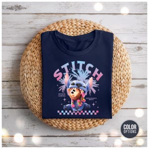Może przedstawiać: Granatowa koszulka z grafiką Stitcha, postaci z Disneya, i słowem "STITCH" powyżej. Projekt zawiera palmy i teksty "Session Surfing" i "Santa Monica Beach". Kolorowy wz&oacute;r w szachownicę znajduje się na dole.