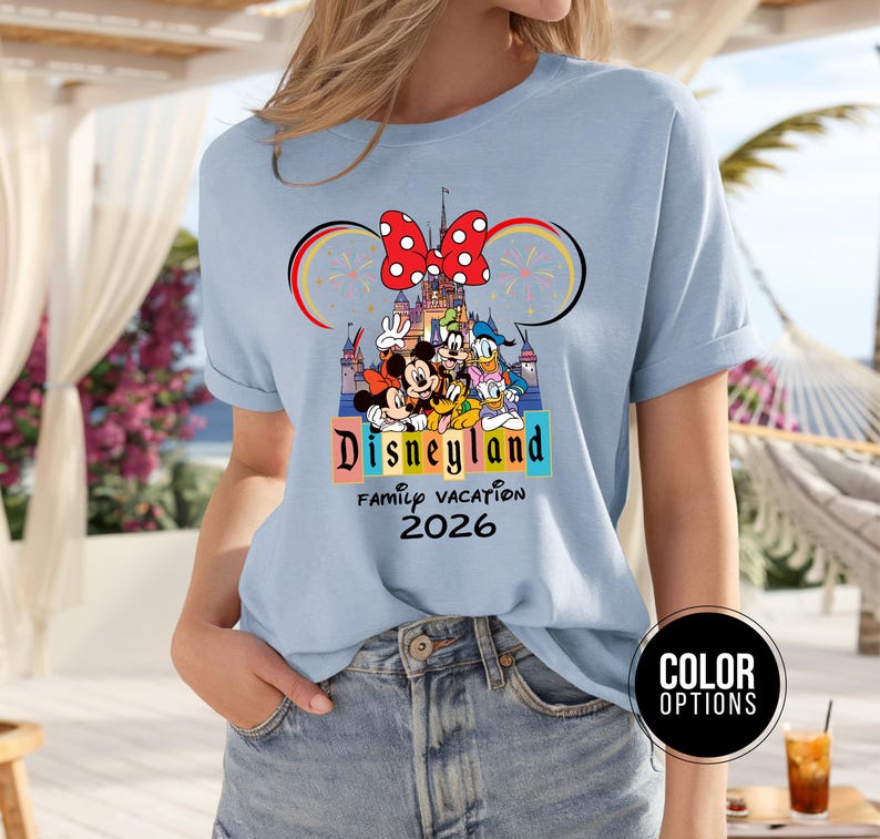 May include: Light blue t-shirt featuring a Disneyland graphic with Mickey Mouse, Minnie Mouse, Donald Duck, and Goofy. The design includes the text "Disneyland Family Vacation 2026" in a colorful font, with a red bow and fireworks.