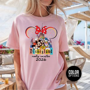 May include: A light pink t-shirt featuring a Disneyland graphic. The graphic includes Mickey and Minnie Mouse, Donald Duck, and Goofy in front of a castle. The shirt also has the text "Disneyland Family Vacation 2026".