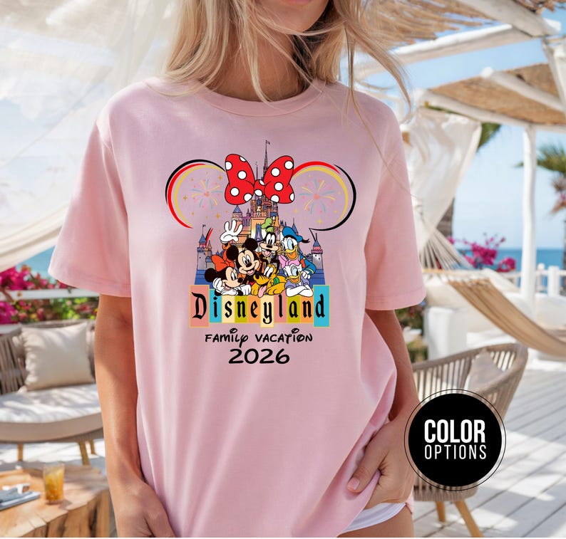 May include: A light pink t-shirt featuring a Disneyland graphic. The graphic includes Mickey and Minnie Mouse, Donald Duck, and Goofy in front of a castle. The shirt also has the text "Disneyland Family Vacation 2026".