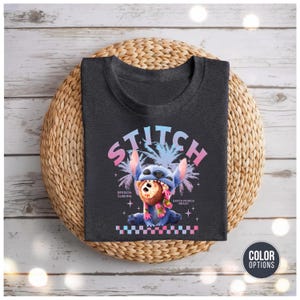 Może przedstawiać: Ciemnoszara koszulka z grafiką Stitcha, niebieskiego kosmity, i napisem "STITCH" powyżej. Projekt zawiera palmy i tekst "Session Surfing Santa Monica Beach". Koszulka leży na plecionej macie.