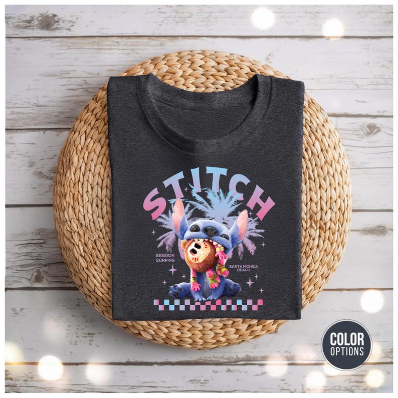 Może przedstawiać: Ciemnoszara koszulka z grafiką Stitcha, niebieskiego kosmity, i napisem "STITCH" powyżej. Projekt zawiera palmy i tekst "Session Surfing Santa Monica Beach". Koszulka leży na plecionej macie.
