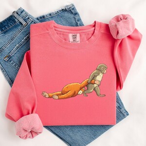 Illustration of a baby macaque named Punch holding a small toy macaque close to his chest, showing an emotional and gentle pose inspired by the viral story of the abandoned monkey, with the larger macaque lying down and the smaller toy being hugged tightly, rendered in a detailed DTF print style with a soft knitted and patch-like stitched effect, featuring clean outlines and layered artwork that gives the design a textured.