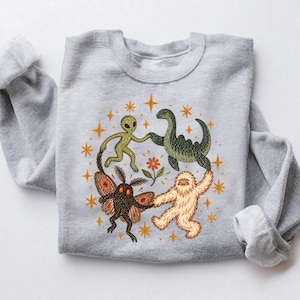 May include: A light gray crewneck sweatshirt featuring a central graphic of an alien, a green dinosaur, a moth, and a white yeti holding hands in a circle around a small orange flower, surrounded by scattered orange four-pointed stars.