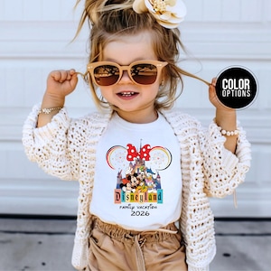 May include: A white t-shirt with a colorful Disneyland graphic, featuring Mickey Mouse, Minnie Mouse, and other characters. The shirt also has the text "Disneyland Family Vacation 2026". The child is wearing sunglasses and a cream-colored cardigan.