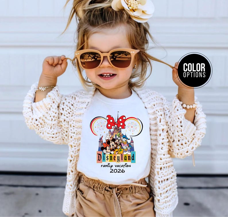May include: A white t-shirt with a colorful Disneyland graphic, featuring Mickey Mouse, Minnie Mouse, and other characters. The shirt also has the text "Disneyland Family Vacation 2026". The child is wearing sunglasses and a cream-colored cardigan.