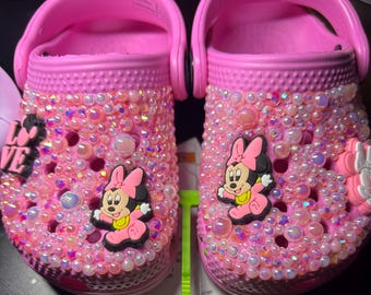 Puede incluir: Un par de zuecos rosa brillante, ricamente adornados con cuentas y abalorios iridiscentes. Cada zapato presenta un dije del personaje de Minnie Mouse y la palabra "Love". Los zuecos están diseñados para tallas infantiles.