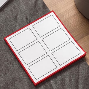 May include: A red-edged notepad with six rectangular white spaces, each outlined in black. The notepad is on a wooden surface, near a laptop and a pen. The notepad is designed for notes or sketches.