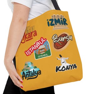 May include: Mustard yellow tote bag with black straps featuring colorful sticker-like graphics of Turkish cities including Istanbul, Izmir, Antalya, and Konya. The bag is carried over the shoulder.