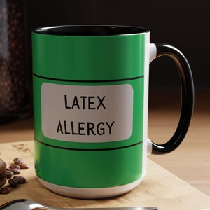 May include: A white ceramic mug with a black handle and rim, featuring a green band with the words "LATEX ALLERGY" in white. The mug sits on a wooden surface with a spoon and a pastry.