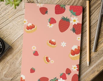 May include: A pink notebook with a pattern of red strawberries, white daisies, and strawberry tarts. A pencil and pen are on the right side of the notebook. The notebook is on a wooden surface.