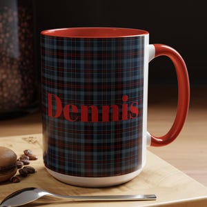 May include: A white ceramic mug with a red interior and handle. The mug features a blue and black plaid pattern with the name "Dennis" in red lettering. The mug is on a wooden surface with a spoon and a pastry.