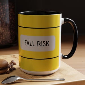 May include: A yellow ceramic mug with a black handle and rim. The mug features the text "FALL RISK" on a white label. A spoon, a macaron, and coffee beans are on a wooden cutting board.