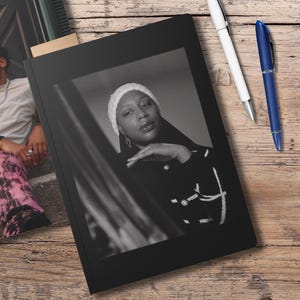 May include: Black journal with a black and white photo of a person wearing a white beanie. A second journal with a photo of a person is visible. Two pens, one white and one blue, are on a wooden surface.