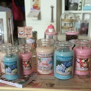 May include: A collection of six glass candle jars with colorful labels. The labels feature floral designs and the text "Yankee Candle". The candles are arranged on a table with a book and other decorative items.