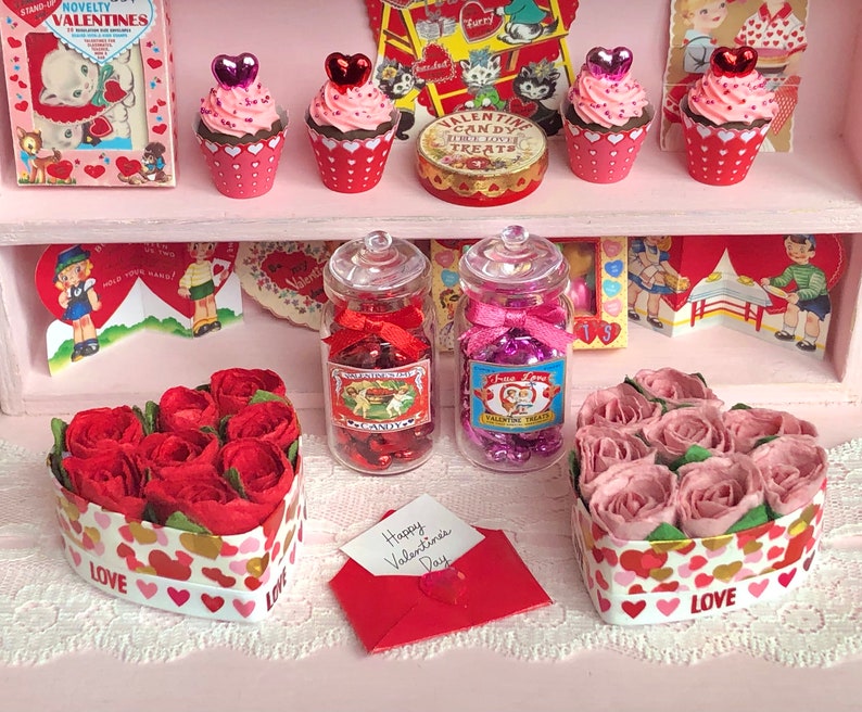 May include: A Valentine's Day display with two heart-shaped boxes filled with red and pink roses, two glass jars filled with candy hearts, and five cupcakes decorated with pink frosting and red hearts.