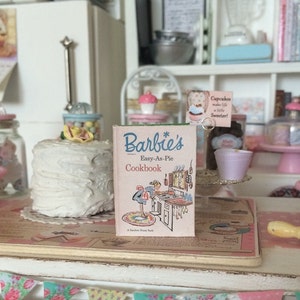 May include: A miniature dollhouse bakery scene with a white cake, jars of candy, and a vintage Barbie cookbook titled "Barbie's Easy-As-Pie Cookbook".