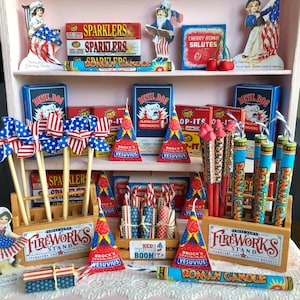 May include: A collection of miniature fireworks, sparklers, and firecrackers in red, white, and blue. The items are arranged on a shelf with a wooden stand and a sign that reads "Fireworks Stand".