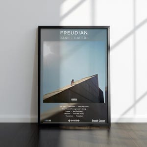 Op de afbeelding: Zwart omkaderde poster met de albumhoes van "Freudian" van Daniel Caesar. De afbeelding toont een persoon op een gebouw tegen een blauwe lucht. De poster bevat de albumtitel, artiestennaam en tracklist.