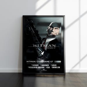 May include: Framed poster featuring a man in a black suit holding a gun, with the title "HITMAN Codename 47" in white text. The poster has a black frame and is displayed against a white wall.