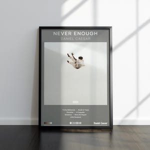 May include: A framed poster with the text "NEVER ENOUGH" and "DANIEL CAESAR" at the top. The poster features a person in mid-air against a light gray background. Below, a list of song titles and the artist's name are displayed.