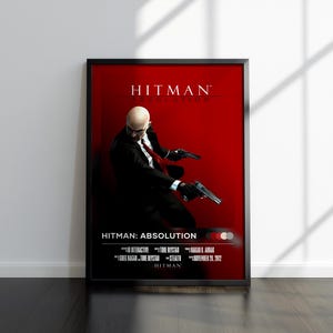 May include: Framed poster for the video game "Hitman: Absolution." The poster features a bald man in a black suit and red tie, holding two guns against a red background. The game title is at the top.