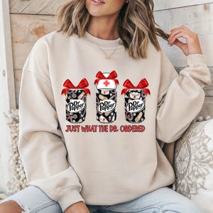Può includere: Felpa beige con tre disegni di lattine di Dr. Pepper con fiocchi rossi. Le lattine presentano grafiche a tema medico, tra cui stetoscopi, bende e un cappello da infermiera. Il testo "JUST WHAT THE DR. ORDERED" è stampato sotto le lattine.