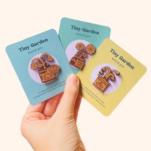Peut inclure: Trois épingles en bois « Tiny Garden » sur des cartes. Chaque épingle est une représentation en bois d'une plante en pot. Les cartes sont bleu clair, vert clair et jaune. Le texte sur les cartes indique « Tiny Garden wood pin ».