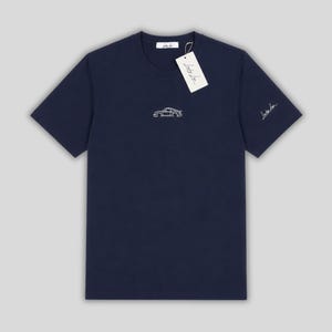 Può includere: T-shirt blu navy con un contorno bianco di un'auto sul davanti. Un'etichetta con il nome del marchio è appesa al colletto. La manica ha una firma bianca. Una maglietta elegante per gli appassionati di auto.