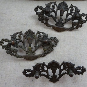 May include: Three antique bronze drawer pulls with ornate, symmetrical designs. Each pull features a central floral element and curved handles, perfect for adding a vintage touch to furniture. The pulls are arranged on a neutral background.