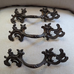 May include: Three antique bronze drawer pulls with ornate, scrolling designs. Each pull features a curved handle and decorative floral accents on the ends. The hardware is likely made of metal and is designed for furniture.