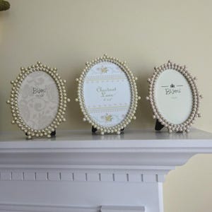 May include: Three oval picture frames with pearl-like bead borders. The frames have black stands and display decorative paper inserts. Two frames have the word "Bijou" printed on them, and one frame has the words "Chestnut Lane" printed on it.