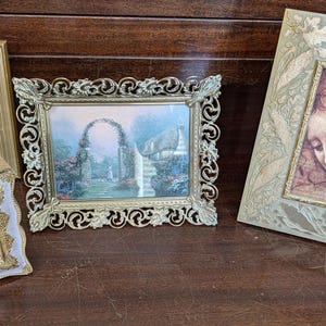 May include: Four decorative picture frames of various sizes and styles are arranged on a dark wooden surface. The frames include a gold-toned frame with a black and white photo, a white and gold frame, and two ornate frames with artwork.