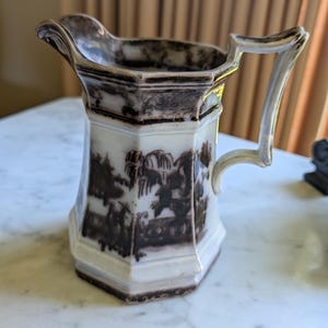 May include: A hexagonal ceramic pitcher featuring a white base with dark brown transferware patterns depicting landscape scenes. The vessel has a prominent pouring spout, a curved handle, and decorative brown trim along the rim and base.