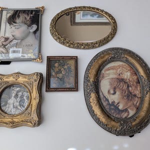 May include: An assortment of antique picture frames and a mirror. The frames vary in shape and size, with ornate gold and silver detailing. One frame holds a black and white photo of a child. Another contains a floral print.