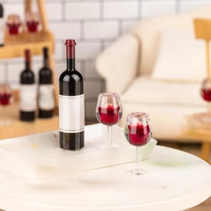 May include: A miniature wine bottle and two wine glasses filled with red liquid. The bottle has a white label and a red cap. The glasses are clear and have a stem. The items are on a white surface.