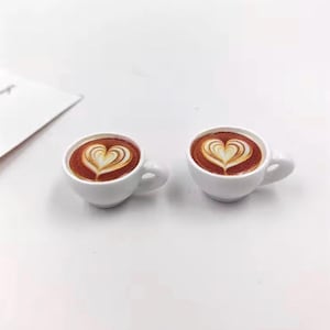 May include: Two small white ceramic coffee cups with heart-shaped latte art. The coffee is a rich brown color, and the heart design is created with a lighter cream color. The cups have small handles and sit on a white surface.