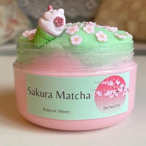 Peut inclure: Un pot rose et vert de slime "Sakura Matcha", orné de petites décorations florales et d'une mini-cornet de glace. L'étiquette indique "Babycat Slimes" et "Do Not Eat".