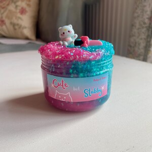 May include: A jar of colorful slime with a small white bear figurine on top. The slime is a mix of pink and blue with glitter. The jar has a label that says "Cute but Stabby."