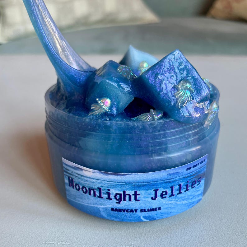 A clear, round plastic container is overflowing with blue sparkly jelly cube slime. The slime is a clear coated slime colorshifting from blue to purple, and has big squishable jelly cubes. It also has tiny opalescent jellyfish charms.