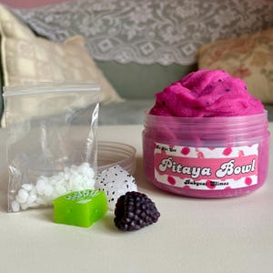 A round transparent container is full of magenta slime with tiny black seeds, resembling a pitaya dragonfruit bowl. on top are beeswax "coconut flakes" and fruit slice charms.