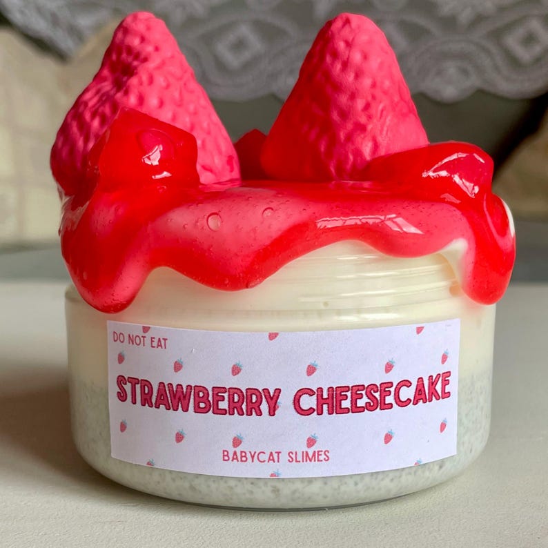 A container labeled "strawberry cheesecake by babycat slimes" is filled with a creme coloured slime with a sand crust, topped with red slime with jelly cubes and strawberry clay pieces.