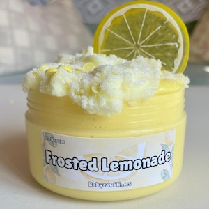 Könnte beinhalten: Ein gelber Schleim in einem durchsichtigen Glas mit einem weißen Etikett, auf dem "Frosted Lemonade" und "Babycat Slimes" steht. Der Schleim ist mit weißen Streuseln und einer gelben Zitronenscheibe bedeckt.