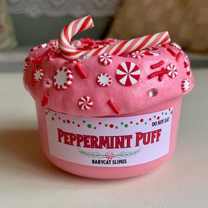 以下が含まれることがあります： 赤と白のキャンディー、スプリンクル、キャンディーケーンで飾られた、ペパーミントパフスライムのピンク色の容器。「Peppermint Puff」と「Do Not Eat」と書かれたラベル。製品はBabycat Slimesからです。