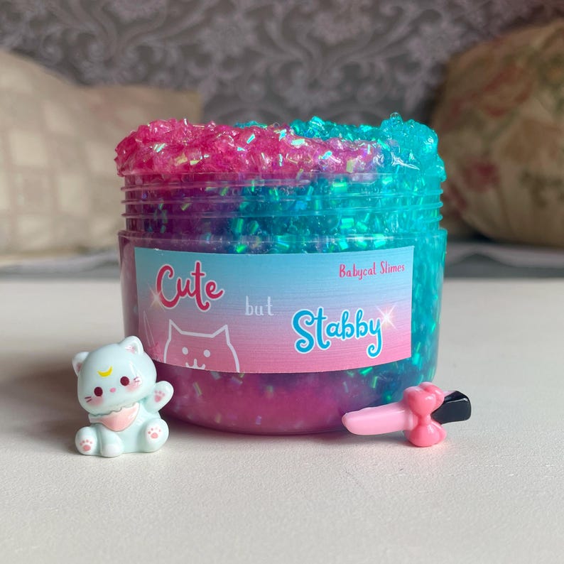 May include: A clear plastic jar filled with pink and blue glitter slime. The jar has a label that reads "Cute but Stabby" with a cat graphic. A small light blue cat figurine and a pink and black toy are next to the jar.