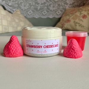 A container labeled "strawberry cheesecake by babycat slimes" is filled with a creme coloured slime with a sand crust, topped with red slime with jelly cubes and strawberry clay pieces.